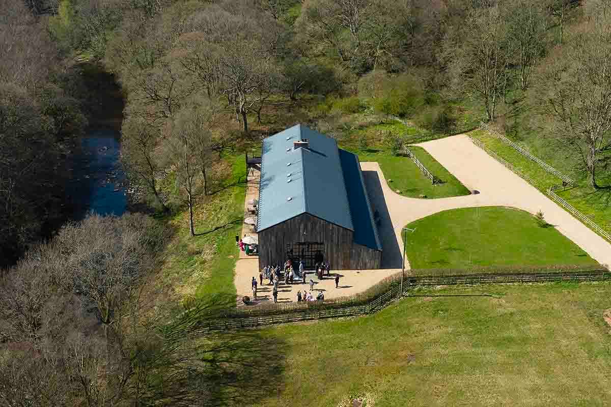 Aerial view of a large barn venue surrounded by lush woodland and manicured grounds on a sunny day.