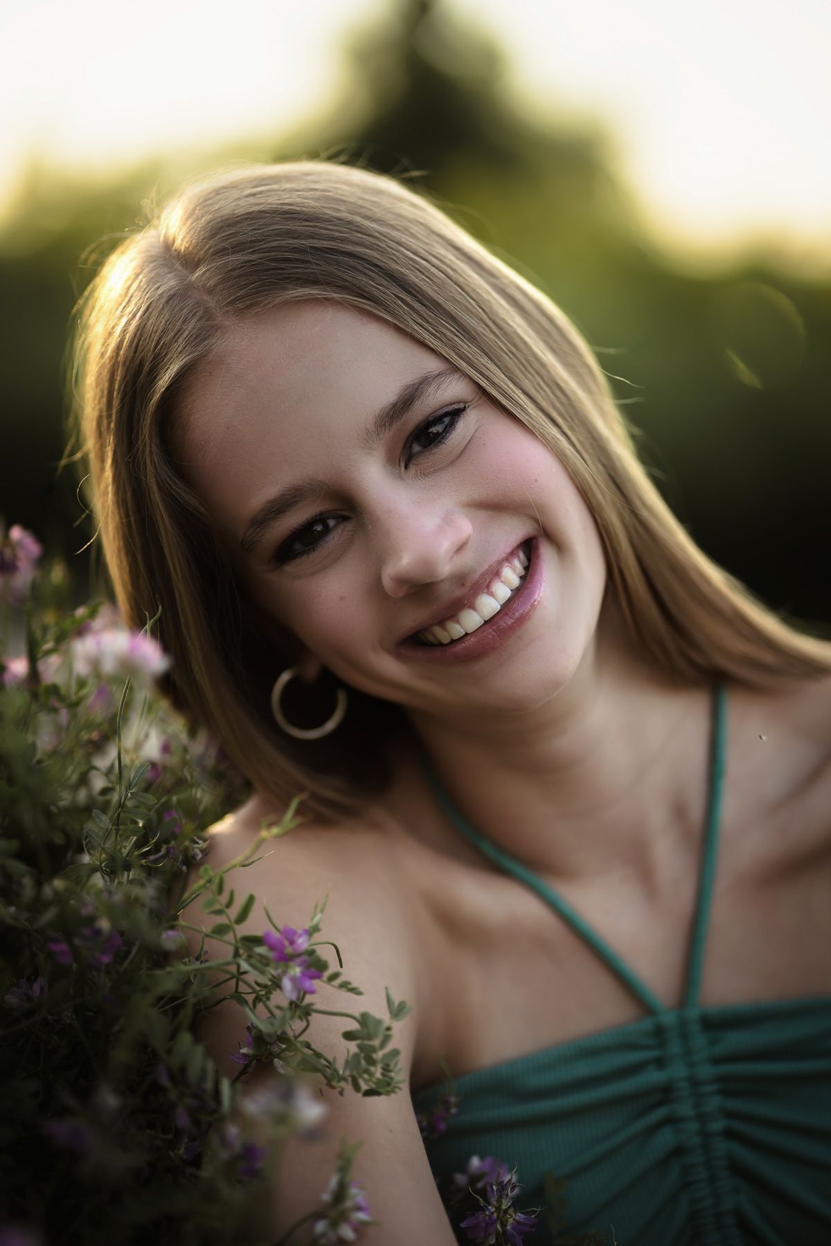 Smiling young blonde woman in green dress holding wildflowers outdoors in golden hour sunlight at senior session