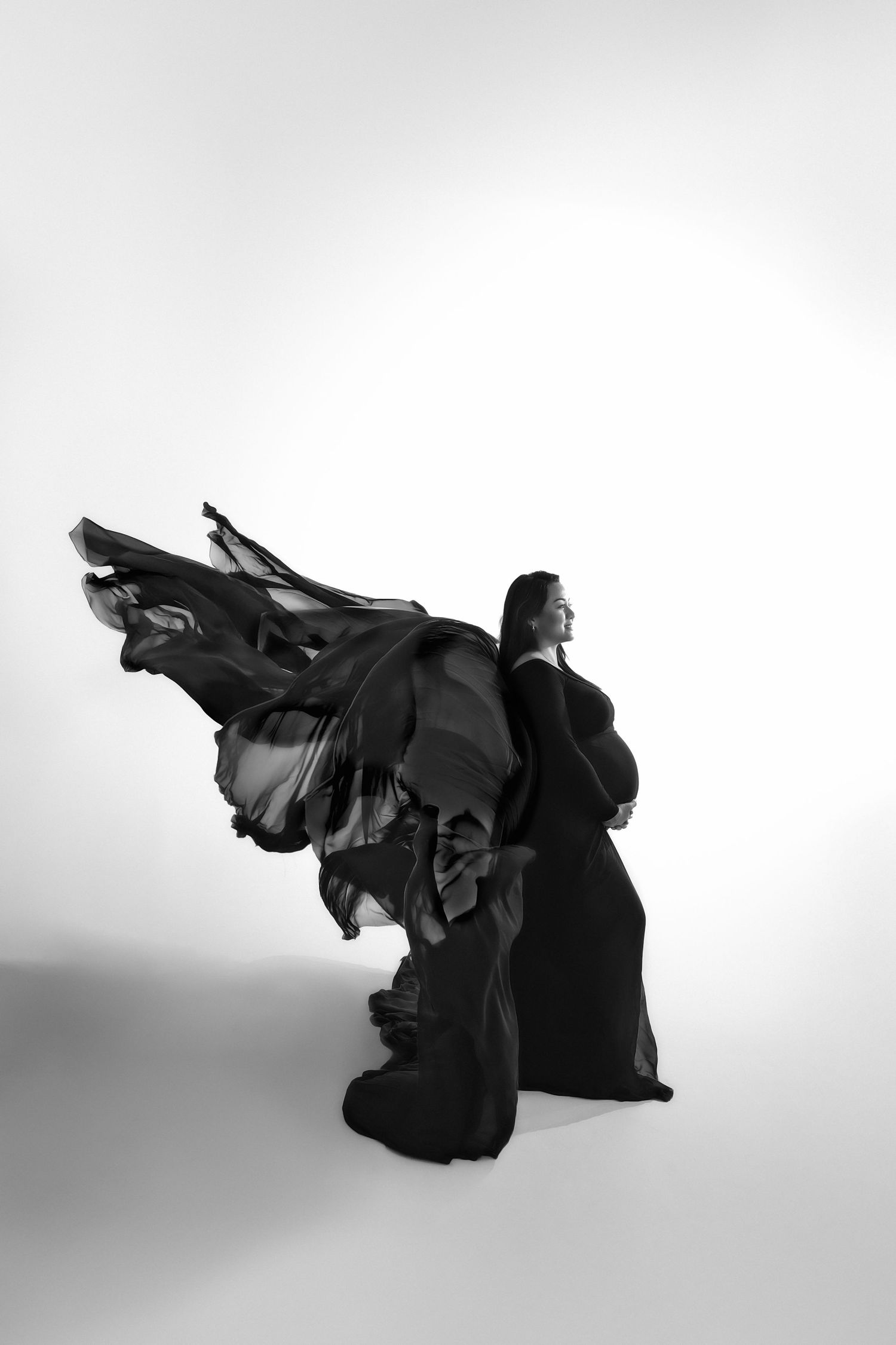 Black and white photo of pregnant woman in flowing dark gown with dramatic fabric billowing behind her in studio setting.