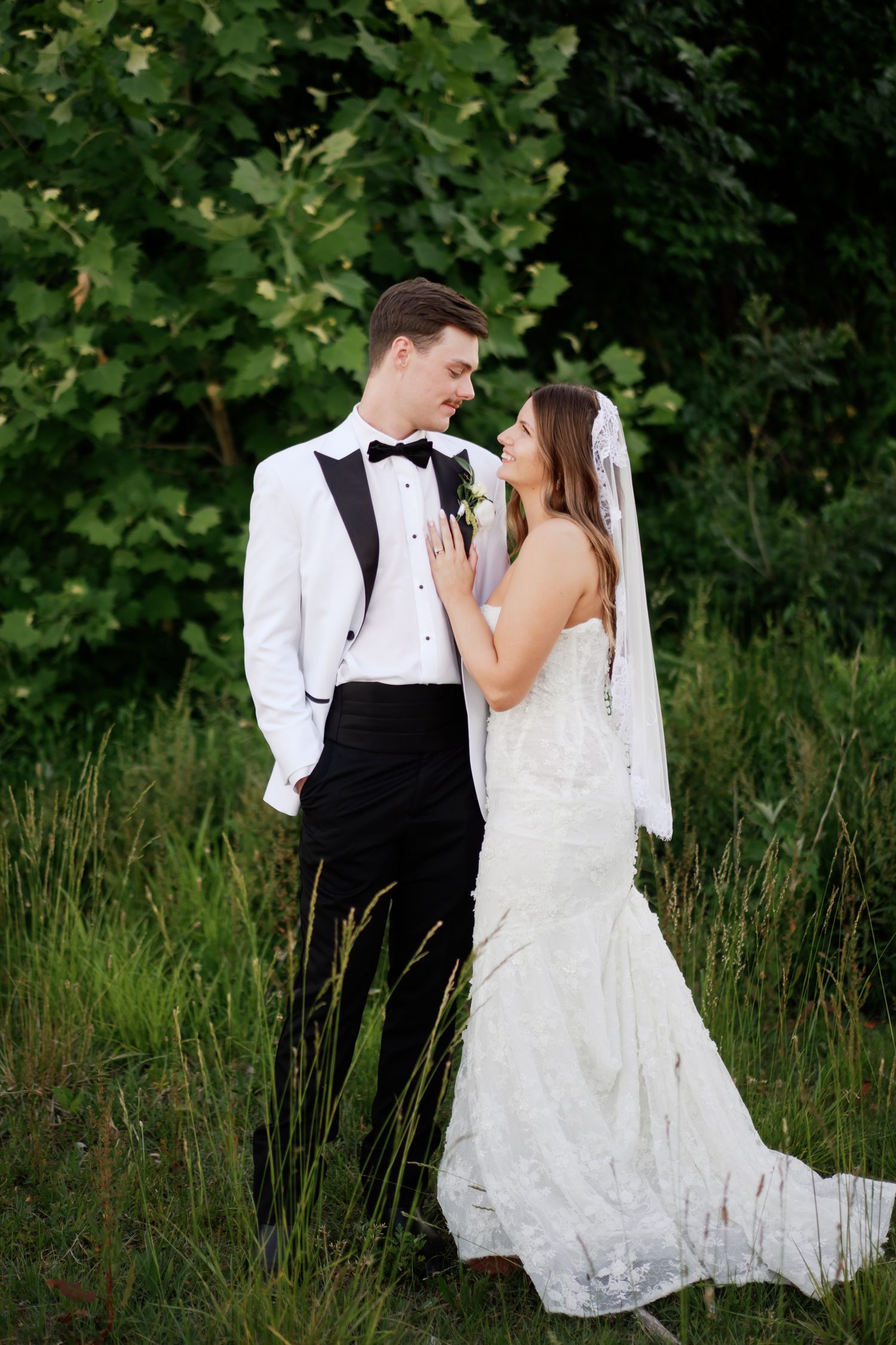 A bride and groom share a romantic moment outdoors, surrounded by lush greenery, as she touches his lapel.