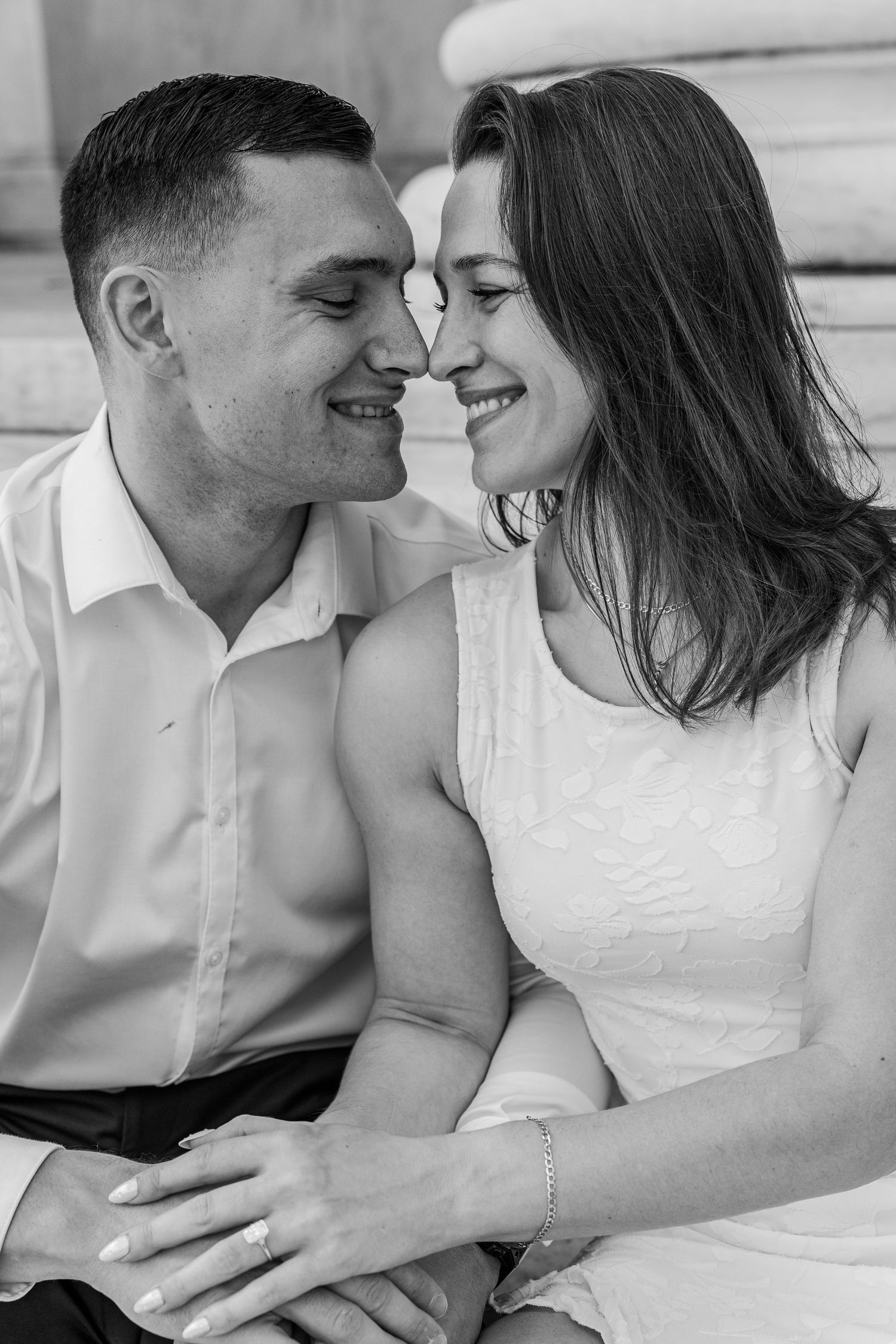 Black and white photo of a romantic couple smiling and touching noses, woman in white dress, man in white shirt.