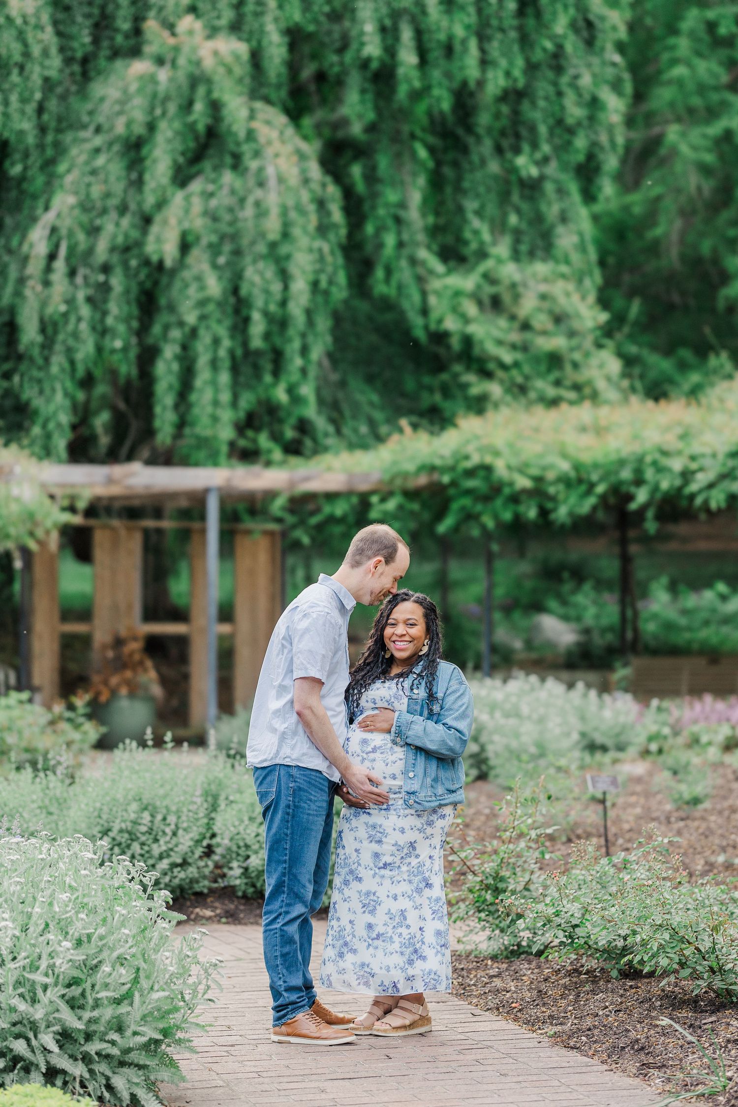 A couple shares a tender moment in a lush garden, the woman in a floral dress showing a baby bump.