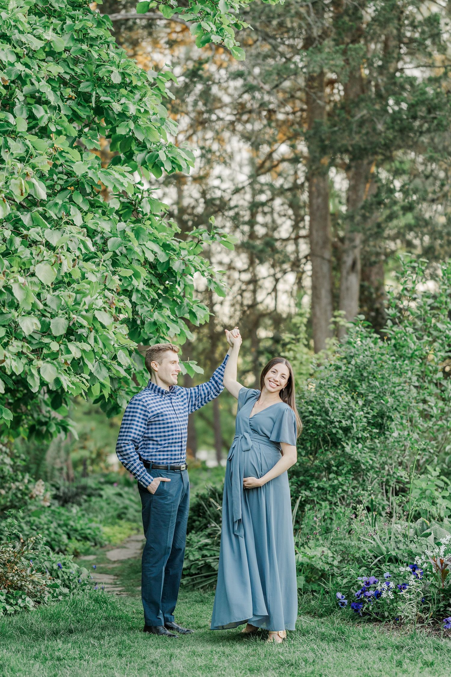 Pregnant couple in a lush green garden, man holding branch above smiling woman in blue dress cradling her baby bump.