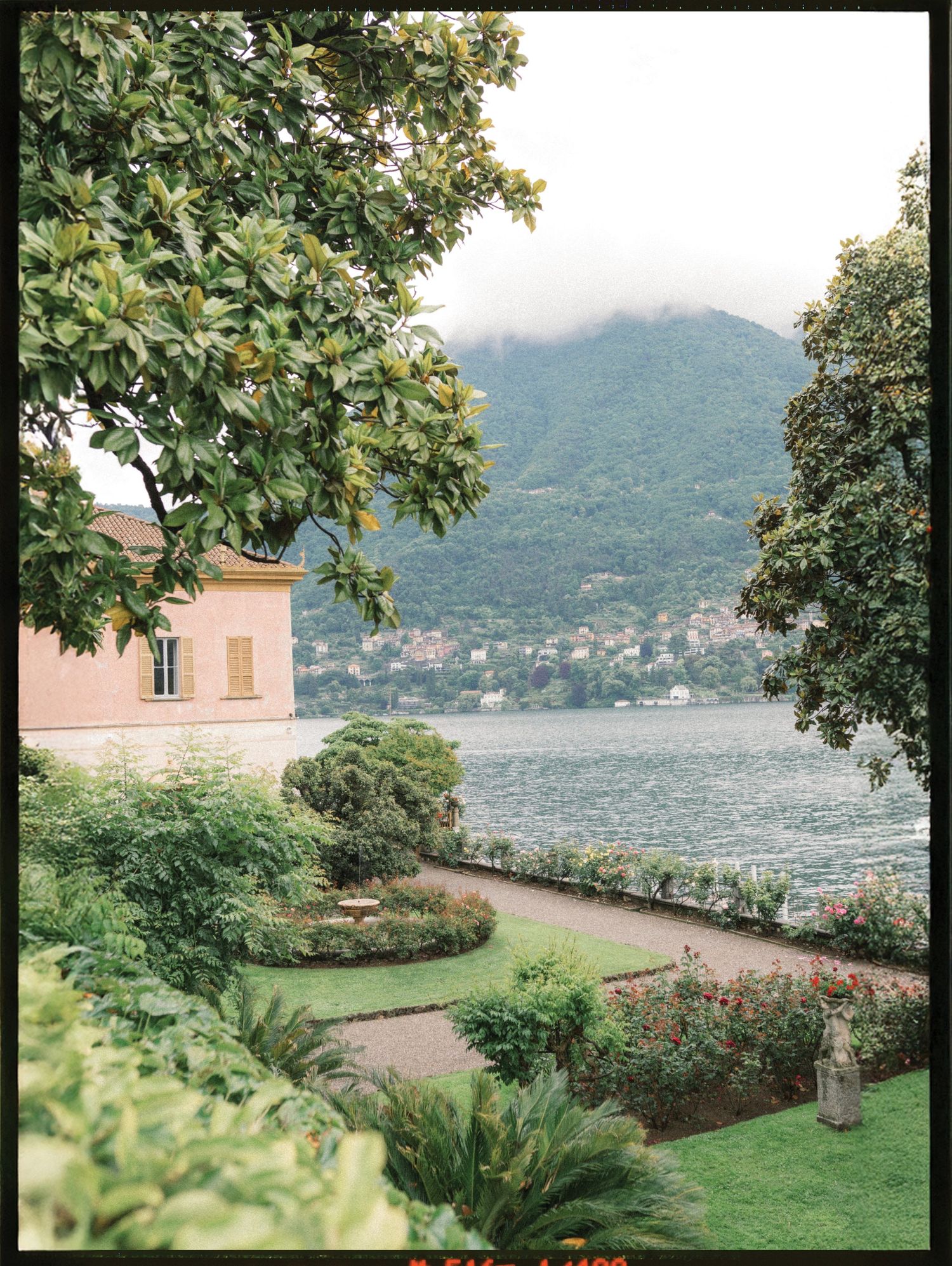 Lush Italian garden with manicured hedges overlooks a serene lake, framed by green mountains and a pink villa.