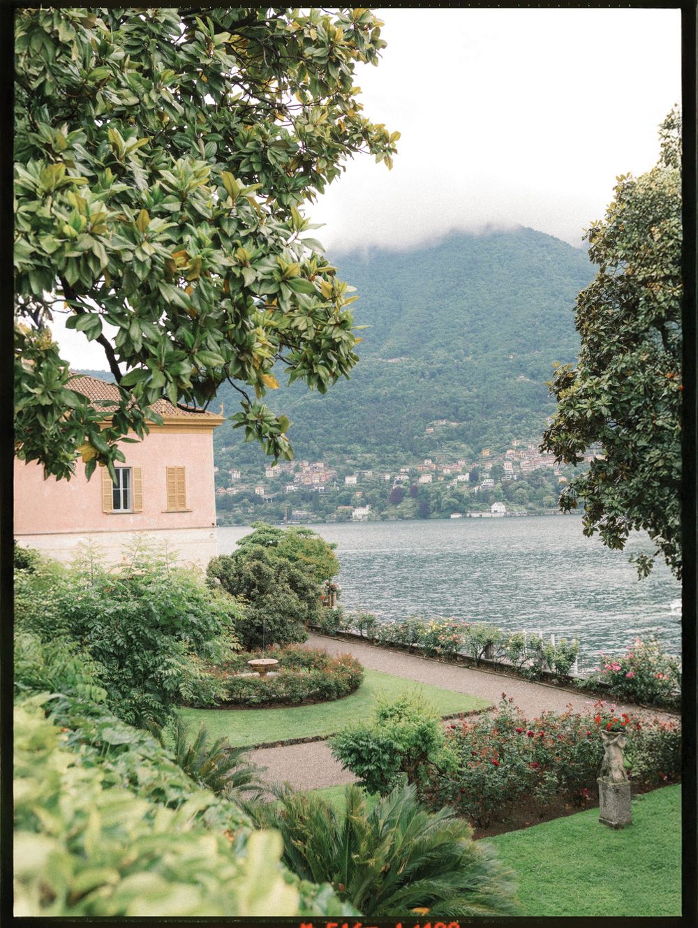 Lush Italian garden with manicured hedges overlooks a serene lake, framed by green mountains and a pink villa.