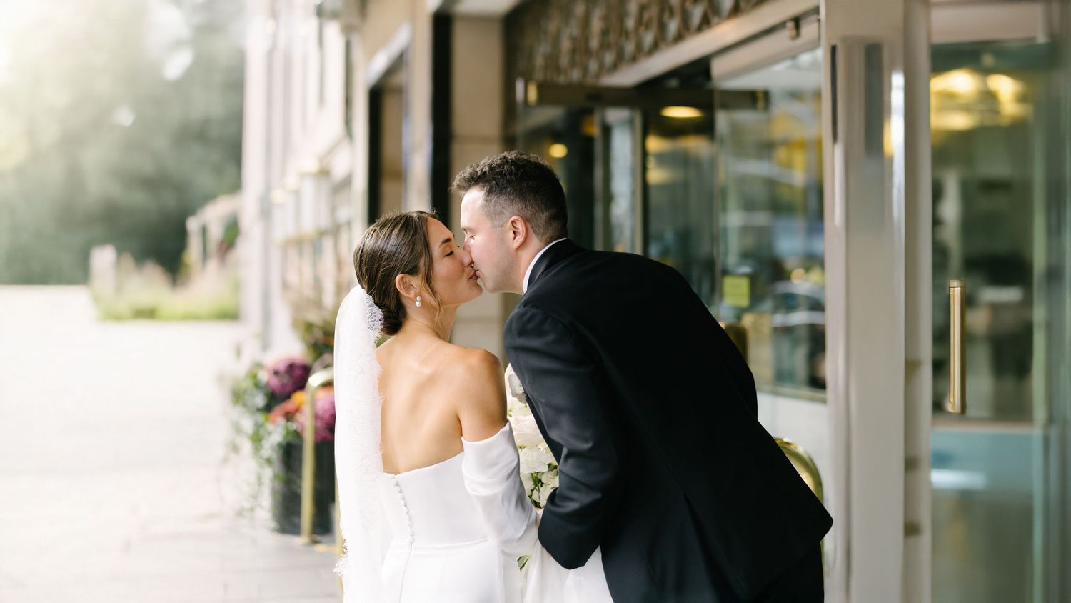 Groom leaning in to kiss the bride outside a glass-fronted building.