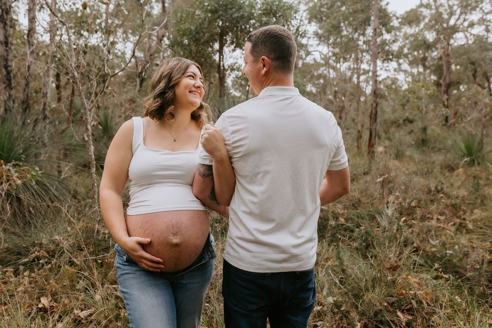 Pregnant woman and partner share a tender moment in a natural bush setting, her baby bump visible in jeans and white top.