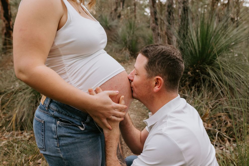 A man kneels to kiss a pregnant woman's belly outdoors among trees during a maternity photo session.