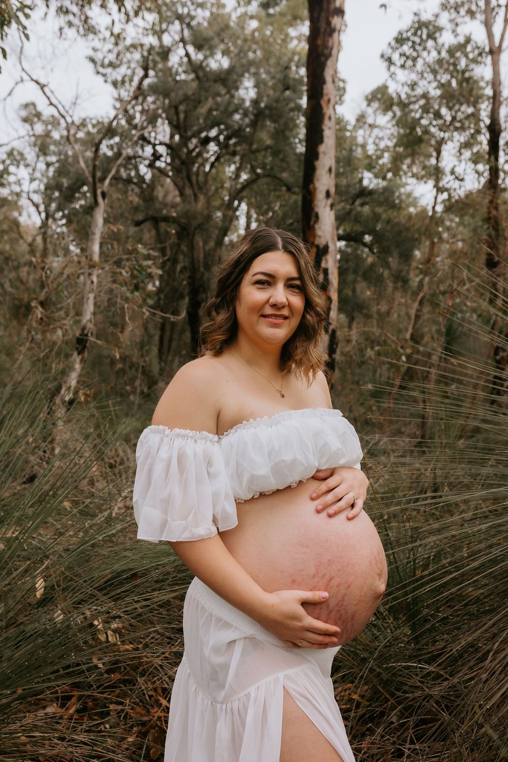 Pregnant woman in white off-shoulder dress cradling baby bump outdoors in Australian bushland setting.