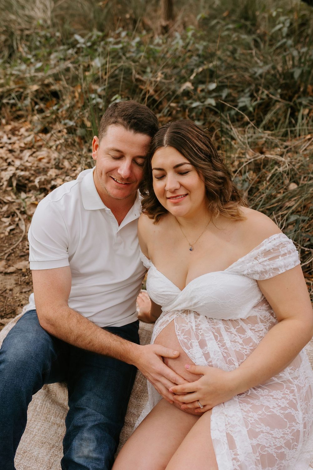 Couple in maternity photoshoot sitting outdoors, man in white shirt and jeans beside pregnant woman in lace dress.