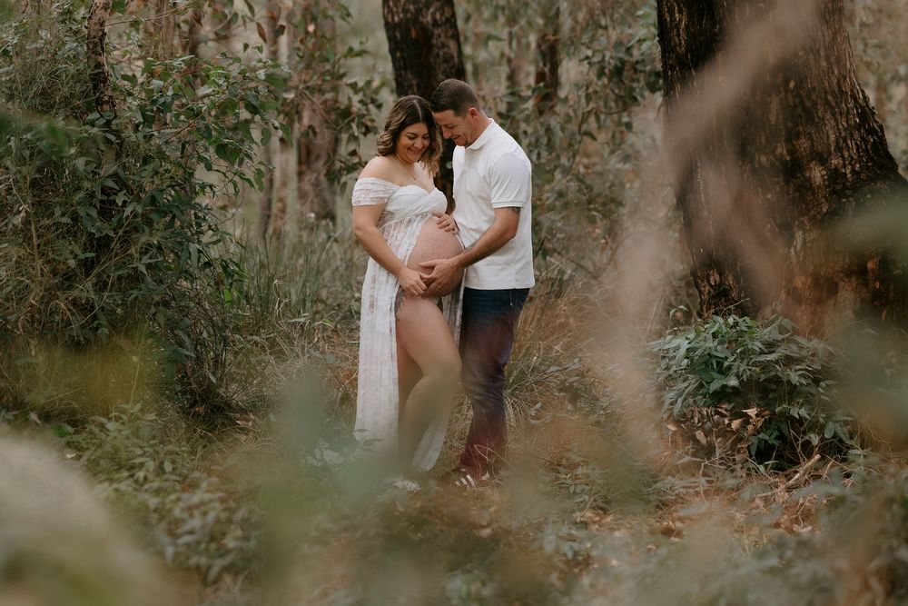 Couple sharing a tender maternity moment in a lush forest, woman in white gown cradling her baby bump.