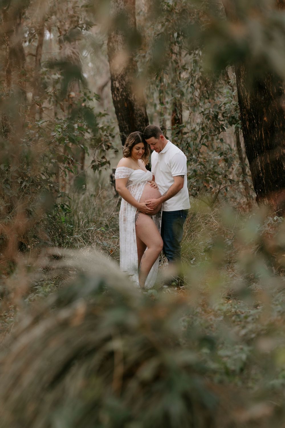 Couple embracing in a lush forest during an outdoor maternity photoshoot, woman in white dress cradling baby bump.