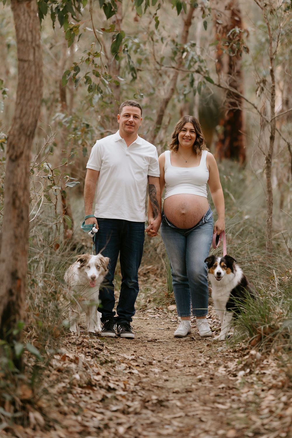 Pregnant couple holding hands on forest path with two Australian Shepherd dogs during maternity photoshoot.
