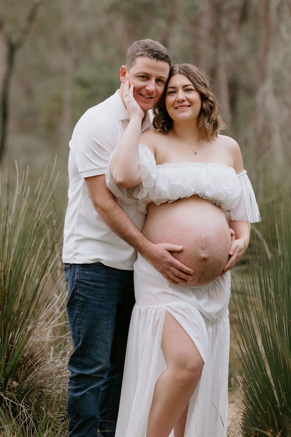 Couple posing for maternity photo shoot outdoors in forest, woman in white off-shoulder dress cradling baby bump.