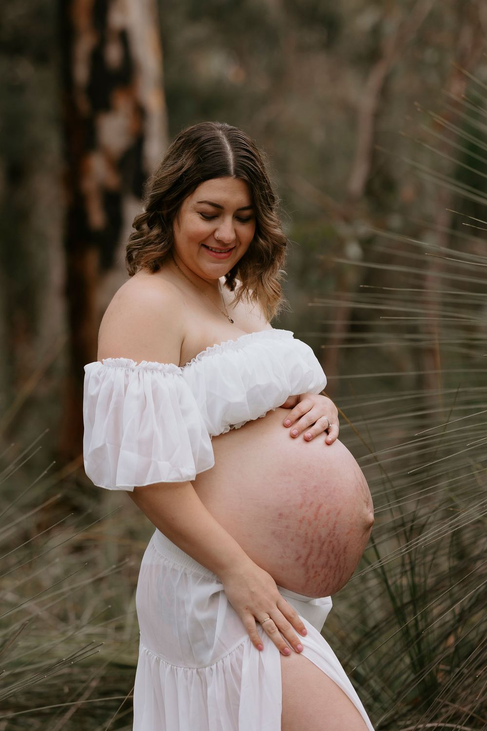 Pregnant woman in white off-shoulder dress cradling her bare baby bump outdoors among tall grass.