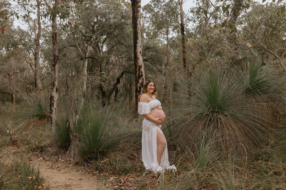 Pregnant woman in white dress posing in Australian bush setting with grass trees and eucalyptus forest backdrop.