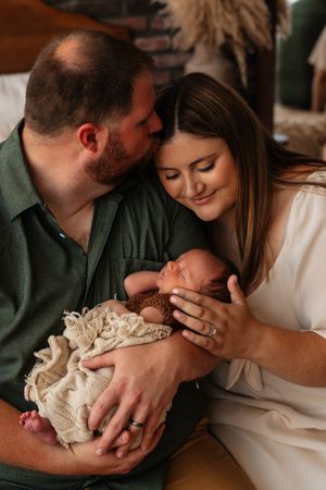 New parents tenderly hold their sleeping newborn baby, with father kissing mother's forehead in a warm, intimate portrait.