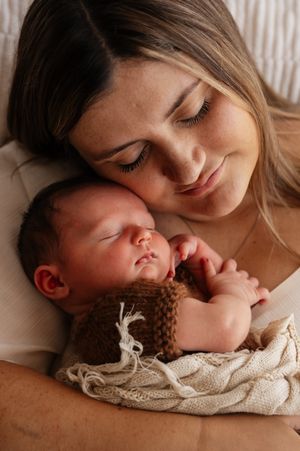 A young mother tenderly cradles her sleeping newborn baby wrapped in a brown knit outfit and cream blanket.