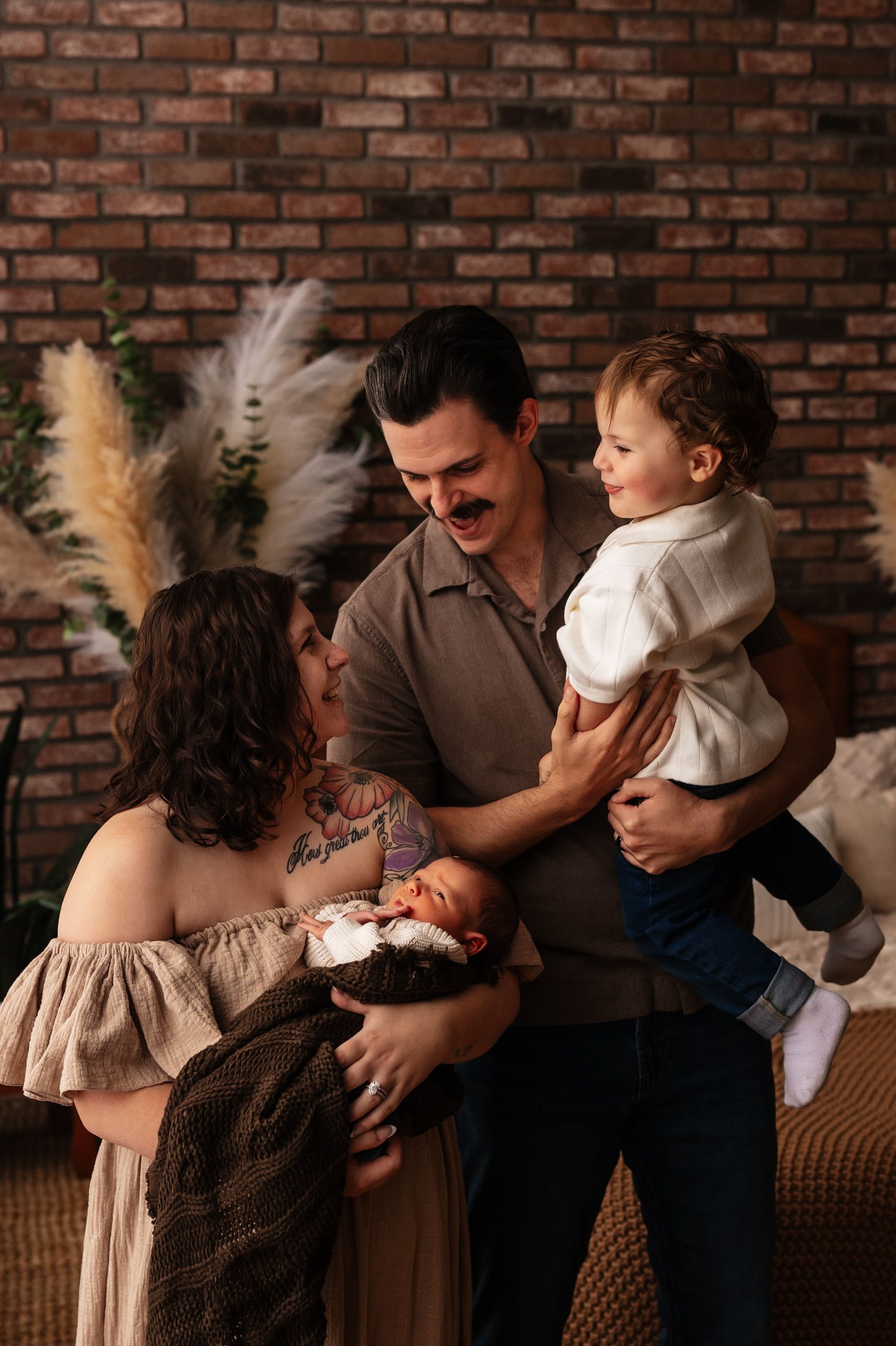 A happy family of four poses in front of a brick wall, parents holding a newborn and toddler in a warm studio setting.