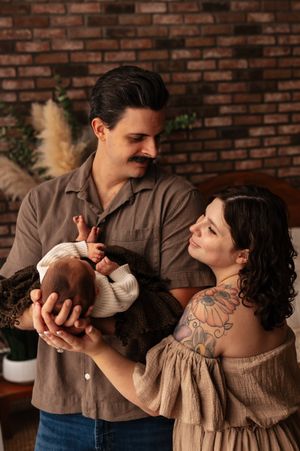 A couple lovingly holds their newborn baby, posing for a portrait against a warm brick wall backdrop.