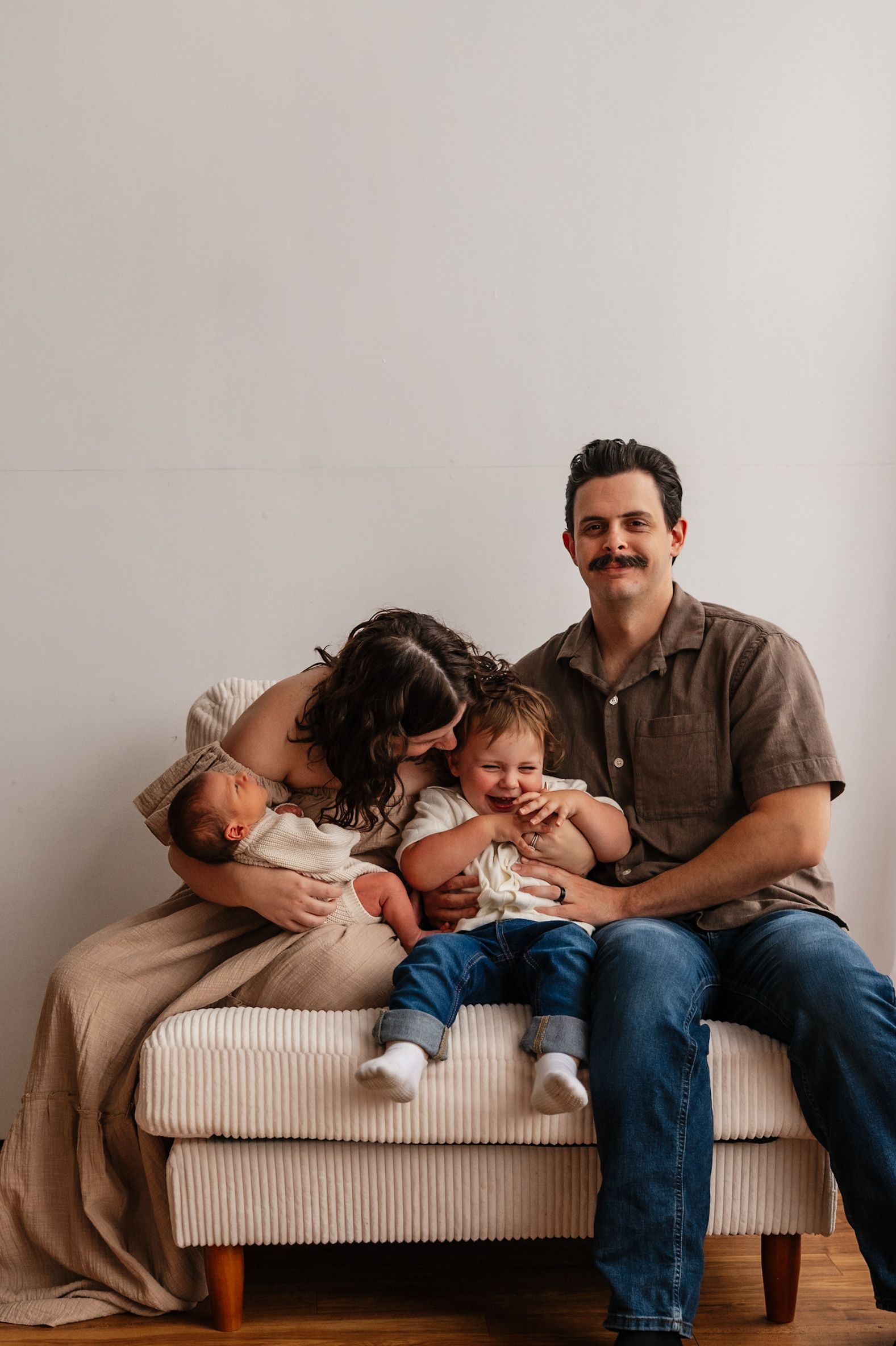 A happy family of four with a mustached dad, mom, toddler, and newborn baby posing together on a cream bench.