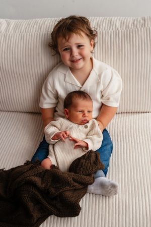 Smiling toddler boy in white shirt holds sleeping newborn baby sibling on a cream couch with brown knit blanket.