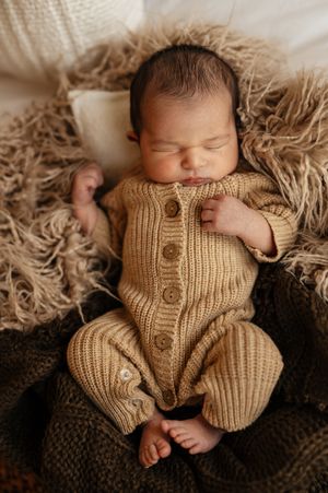 Sleeping newborn baby in a tan knit romper lying on a fluffy beige blanket in a cozy nest setup.