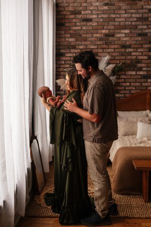 A couple in a cozy bedroom gazes lovingly at their newborn baby near a window with sheer curtains and brick wall.