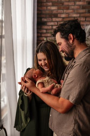 Happy couple smiling at their newborn baby near a window with brick wall background.