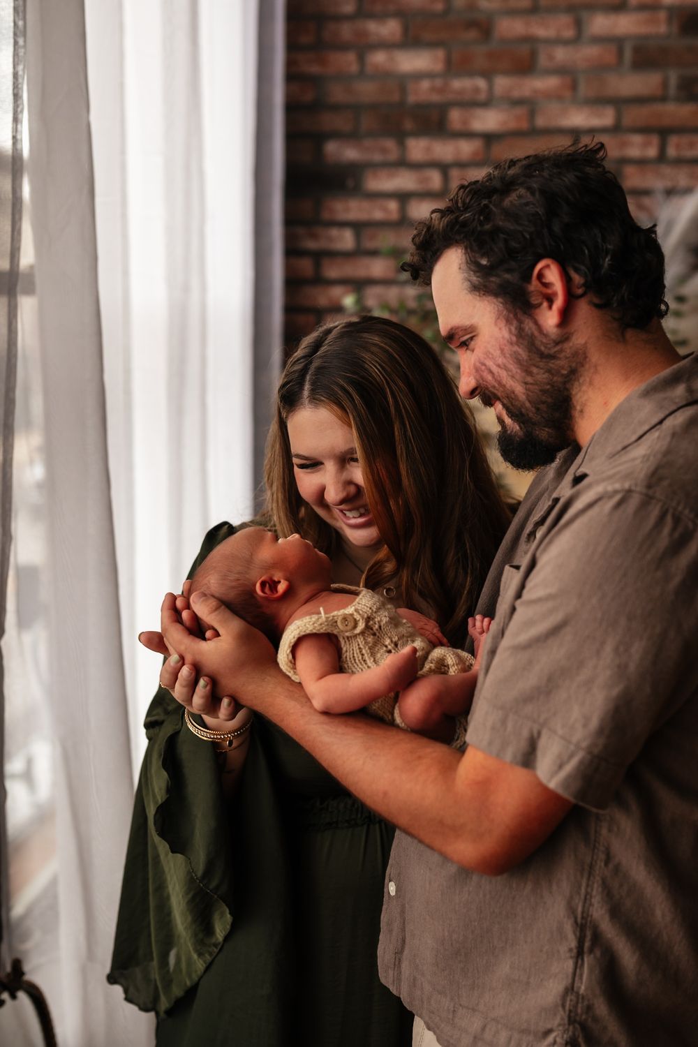 Happy couple smiling at their newborn baby near a window with brick wall background.