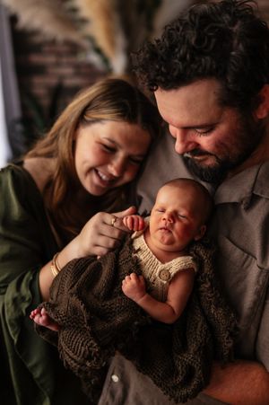 Happy parents smiling at their sleeping newborn baby wrapped in a brown knit blanket during a family portrait session.
