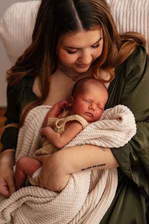 A young mother in a green dress tenderly cradles her sleeping newborn wrapped in a soft cream knit blanket.
