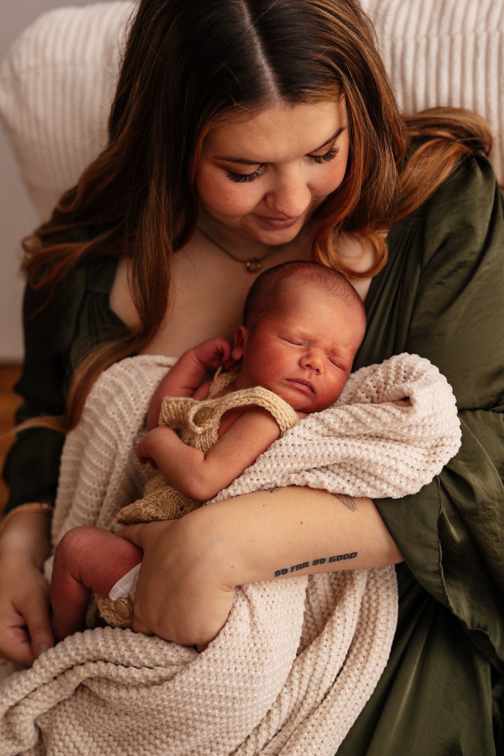 A young mother in a green dress tenderly cradles her sleeping newborn wrapped in a soft cream knit blanket.