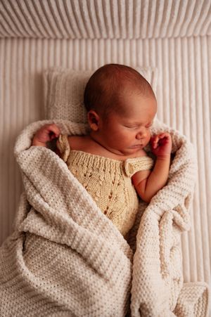 Peaceful newborn baby sleeping wrapped in a cream knit blanket wearing a yellow knit romper on a striped background.