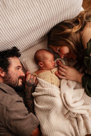 New parents lying on bed gazing lovingly at their sleeping newborn baby wrapped in a cream knit blanket.