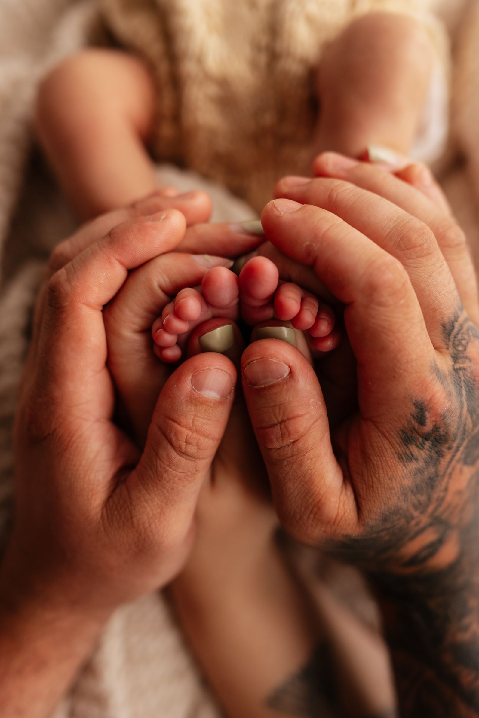 Tattooed adult hands gently cradling tiny newborn baby feet in a tender close-up portrait.