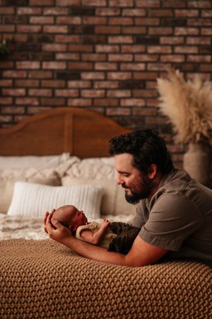 A bearded father lies on a bed holding his newborn baby, gazing lovingly at the infant against a brick wall backdrop.