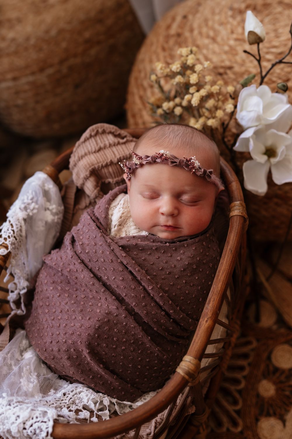 Newborn baby swaddled in brown wrap with floral headband sleeping in wicker basket with white flowers.