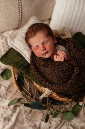 Sleeping newborn baby wrapped in dark green knit blanket nestled in wicker basket with eucalyptus leaves.