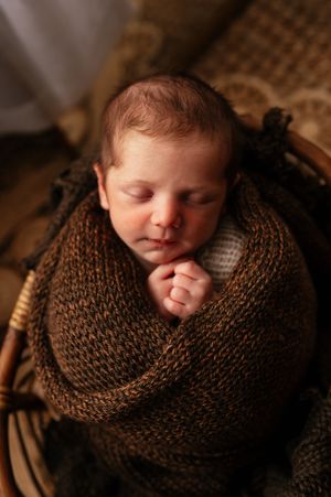 Sleeping newborn baby swaddled in a brown knit wrap, nestled in a wicker basket.