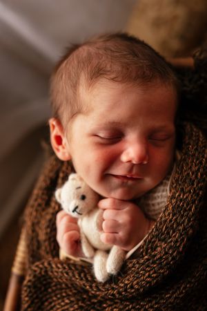 Peaceful newborn baby sleeping while clutching a tiny stuffed bear wrapped in a knit blanket.