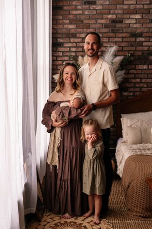 A smiling family of four poses indoors, with mom holding a newborn, dad standing beside, and a young toddler girl in front.
