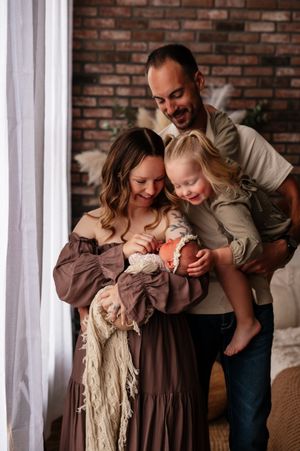 Family of four posing indoors, mother in brown dress holding newborn, father carrying toddler girl, brick wall background.