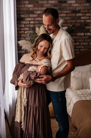 A couple smiles lovingly at their newborn baby in a warmly decorated studio with brick walls and boho decor.