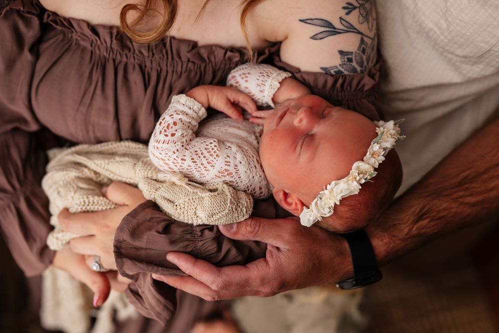 Parents cradling a sleeping newborn baby dressed in a cream knit outfit and floral headband.