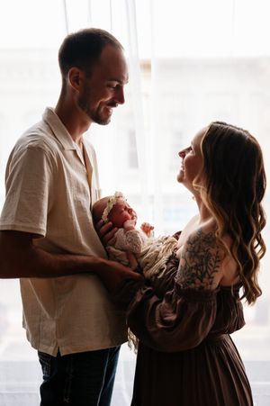 New parents smiling at each other while holding their newborn baby wrapped in white lace near a bright window.
