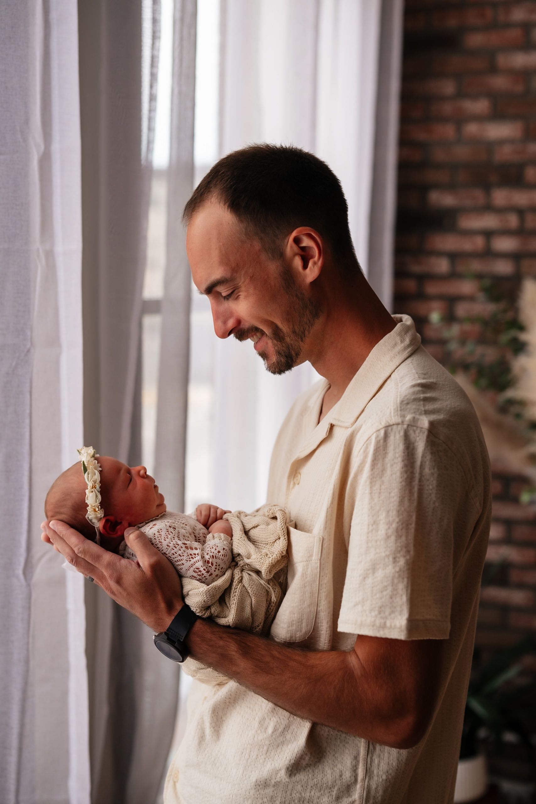 Smiling father holding his newborn baby near a bright window in a cozy brick-walled room.