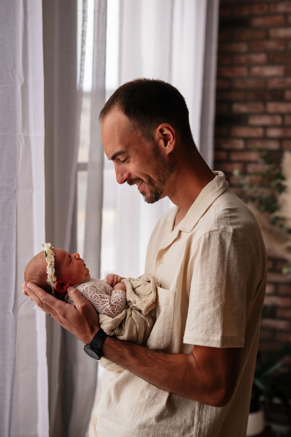 Smiling father holding his newborn baby near a bright window in a cozy brick-walled room.