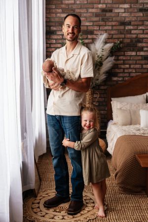 Father holding newborn baby while toddler daughter hugs his legs, smiling in a cozy bedroom setting.