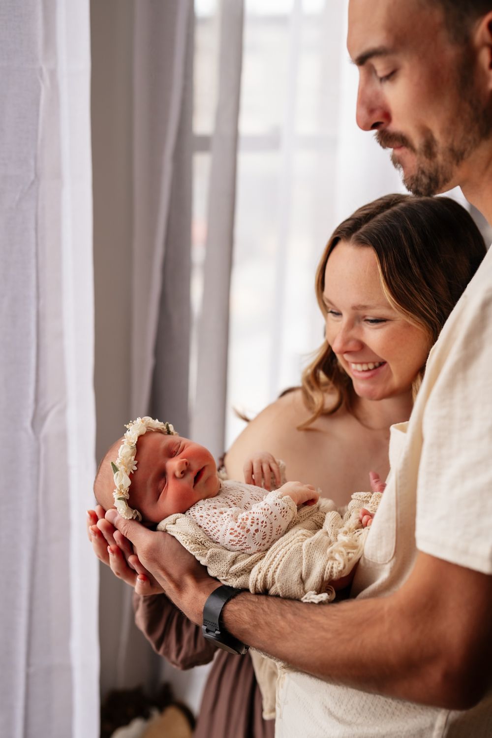Happy parents smiling at their newborn baby girl in a floral headband near a bright window.